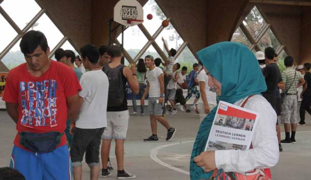 Eine Gruppe junger Leute in einer Jugendfreizeiteinrichtung, einige spielen Basketball, eine Frau hält ein „Deutsch lernen“ Buch.