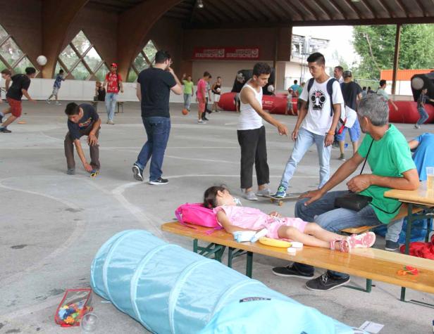 Eine schlafende Junge auf einer Bank, während im Hintergrund andere Menschen spielen.