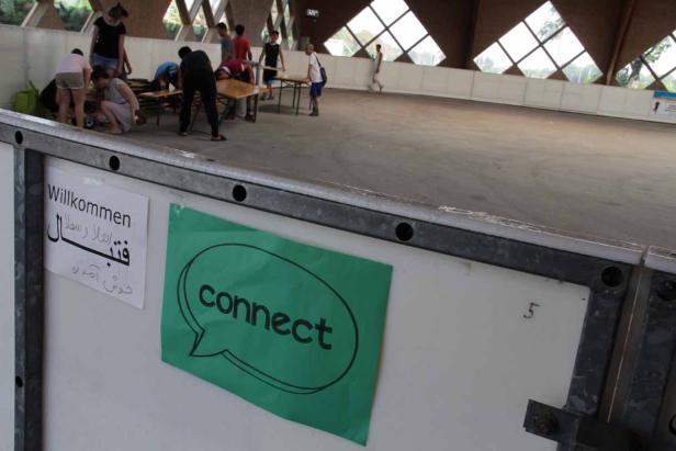 In einer Halle bereiten Menschen Tische vor, ein Schild heißt „Willkommen“.