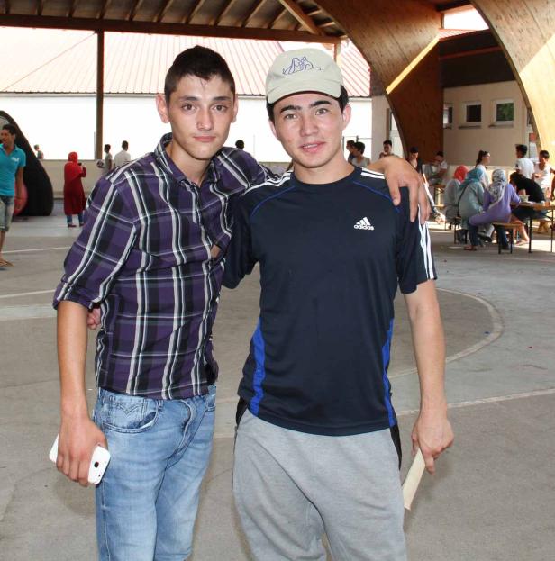 Zwei junge Männer stehen im Freien, einer mit kariertem Hemd und der andere mit Sportbekleidung.