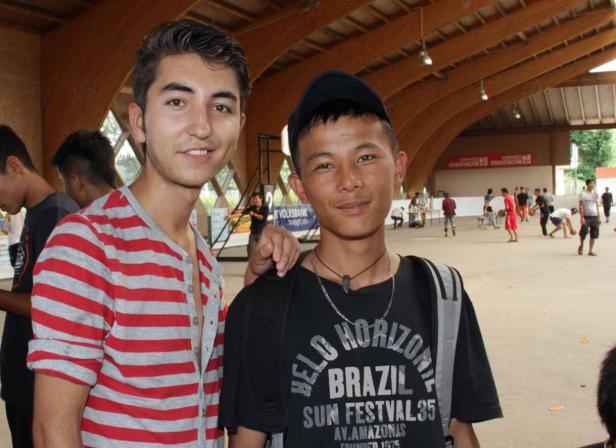 Zwei junge Männer posieren vor einer Gruppe Basketball spielender Personen in einer Halle.