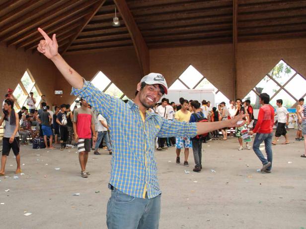 Ein Mann mit Kappe und kariertem Hemd steht mit ausgestreckten Armen in einer Halle voller Menschen.