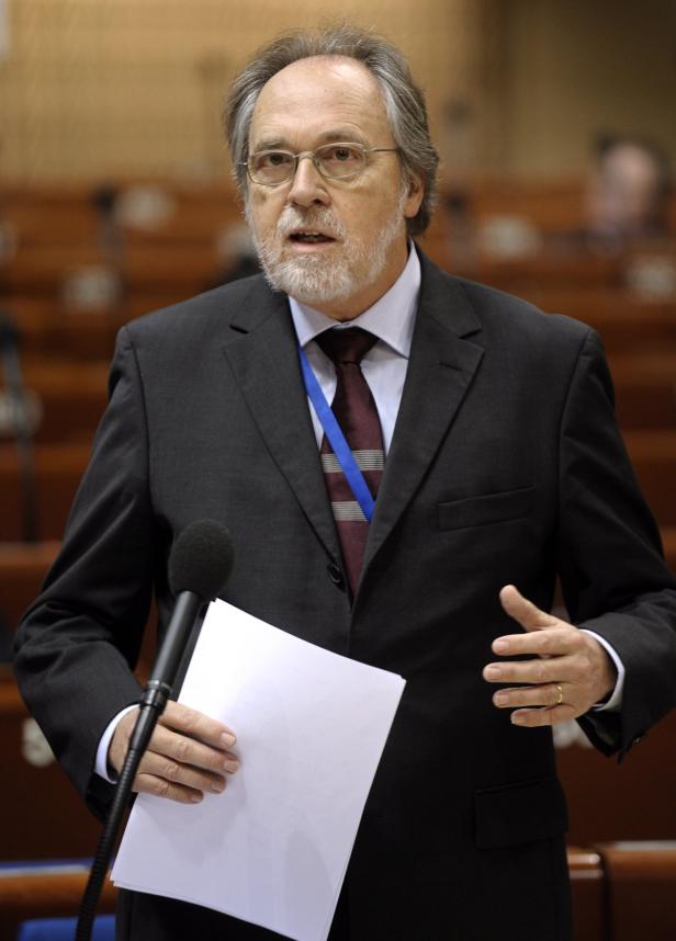 Ein Mann mit Brille und Anzug spricht am Mikrofon und hält ein Blatt Papier.