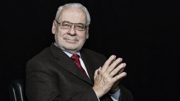 Ein älterer Mann mit Brille und Anzug sitzt vor einem schwarzen Hintergrund.