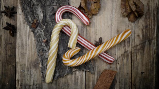 Drei Zuckerstangen liegen auf einem Holztisch mit Sternanis und Tannenzapfen.