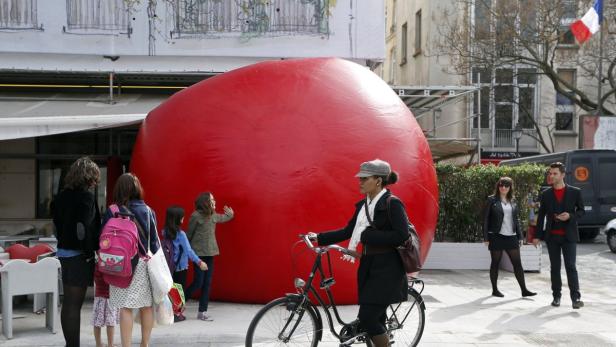 Eine Frau mit Fahrrad steht vor einer großen, roten, aufgeblasenen Skulptur in einer Stadt.