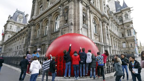 Eine Gruppe von Kindern interagiert mit einer großen, roten Kugel vor einem Gebäude in Paris.