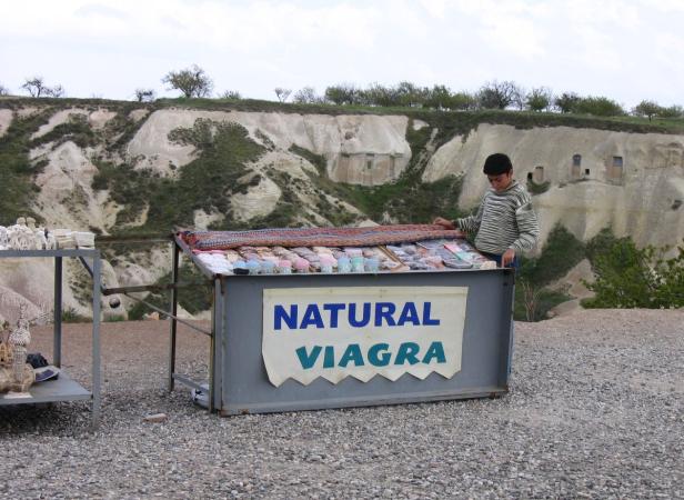 Ein Junge steht an einem Verkaufsstand mit der Aufschrift „Natural Viagra“ in einer Hügellandschaft.