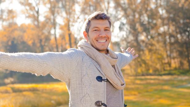 Ein lächelnder Mann mit Schal und Strickjacke steht im Herbstwald und streckt die Arme aus.