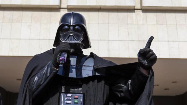 Eine Person im Darth Vader Kostüm spricht in ein Mikrofon und hebt den Zeigefinger.
