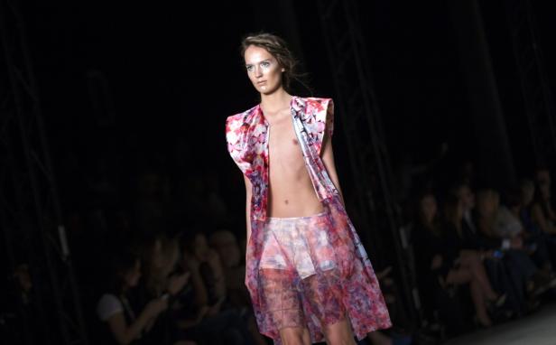 Ein Model präsentiert ein transparentes, buntes Kleidungsstück auf einem Laufsteg.