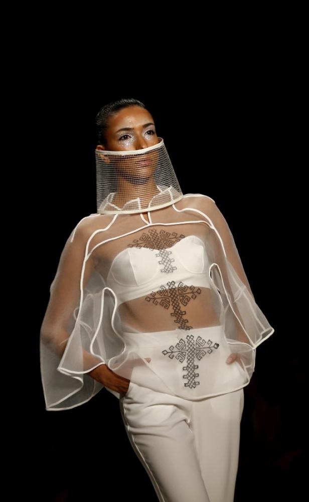 Eine Frau präsentiert ein weißes Ensemble mit transparentem Überwurf und Kopfbedeckung auf dem Laufsteg.