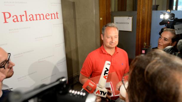 Ein Mann in rotem Hemd steht vor Mikrofonen im österreichischen Parlament.