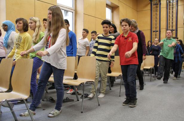Eine Gruppe von Schülern macht Übungen mit Stühlen in einem Raum.