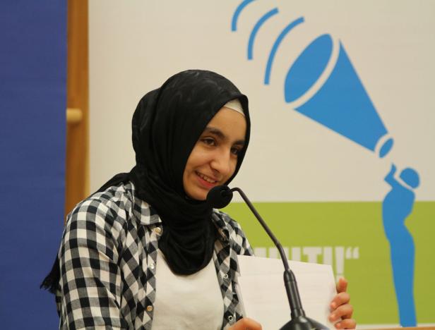 Eine junge Frau mit Hijab spricht bei einer Veranstaltung in ein Mikrofon.