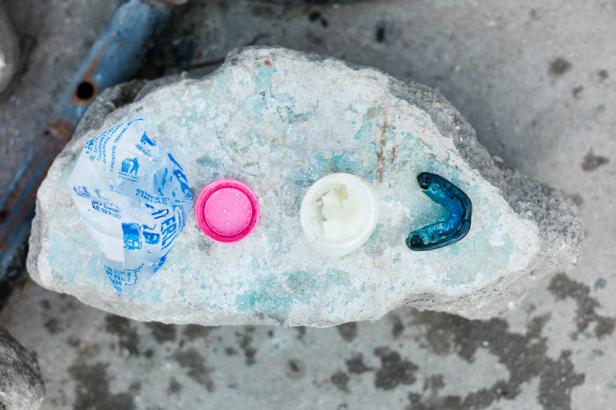 Ein blauer Mundschutz, eine Dose Vaseline, eine rosa Kappe und eine Tüte Wasser liegen auf einem Stein.