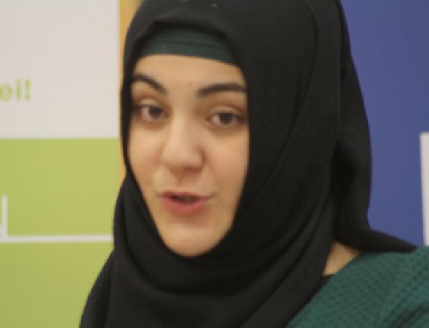 Eine Frau mit dunklen Haaren und einem schwarzen Hidschab spricht.