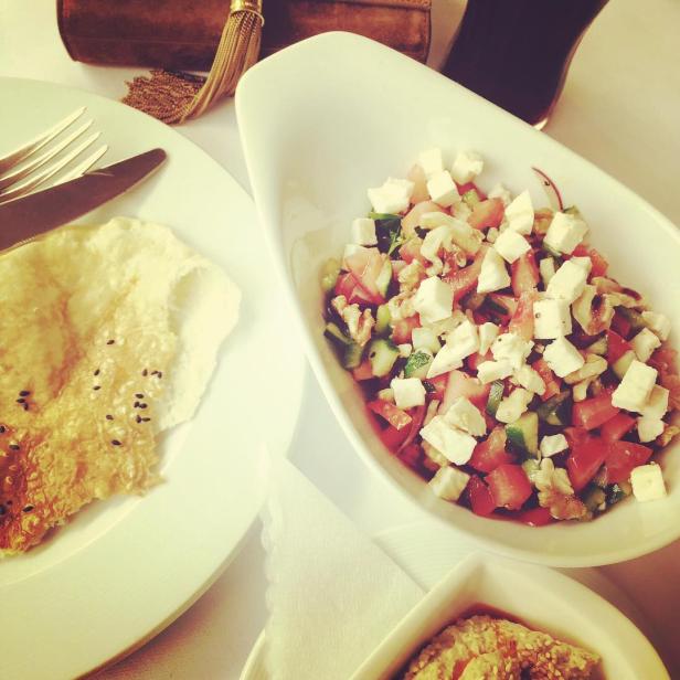 Ein Teller mit Brot, ein Salat mit Tomaten und Feta, und eine Schale mit Hummus stehen auf einem Tisch.