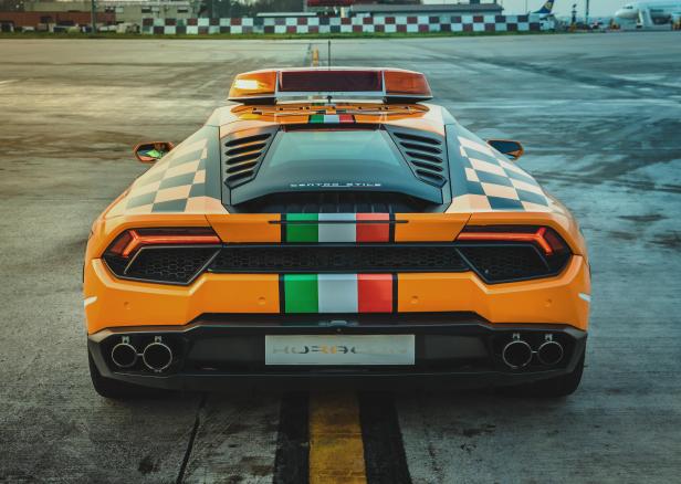 Ein orangefarbener Lamborghini Huracán mit Schachbrettmuster und italienischen Flaggenstreifen auf einem Flugfeld.