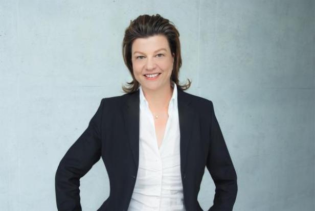 Eine lächelnde Frau in Blazer und Bluse vor einem hellen Hintergrund.