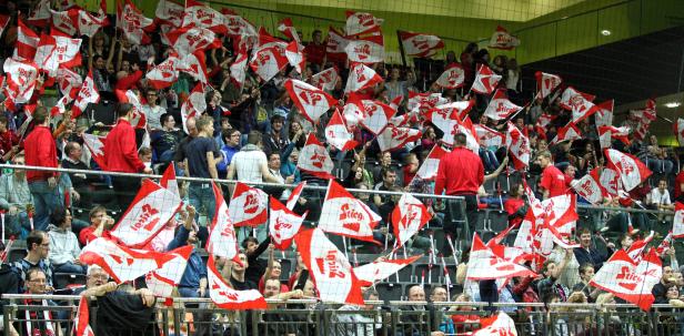 Zuschauer schwenken rot-weiße Fahnen mit der Aufschrift „Stiegl“ bei einem Handballspiel.