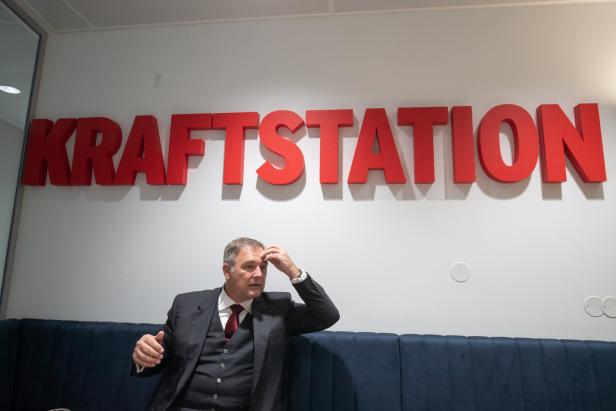 Ein Mann im Anzug sitzt vor einem großen roten „KRAFTSTATION“-Schriftzug und hält sich die Stirn.