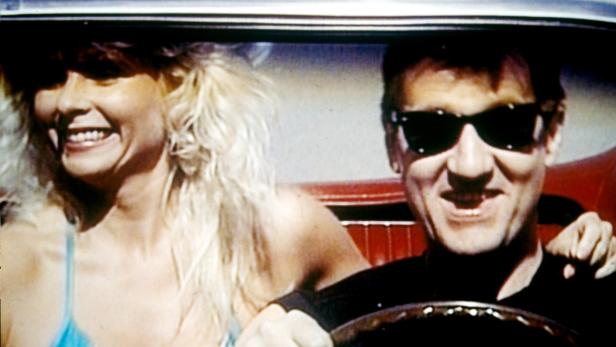 Ein Mann mit Sonnenbrille fährt ein Auto mit einer blonden Frau auf dem Beifahrersitz.