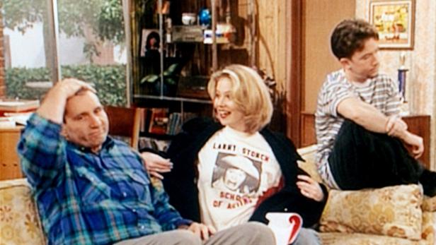 Drei Personen sitzen auf einem Sofa, eine Frau trägt ein T-Shirt von Larry Storch.