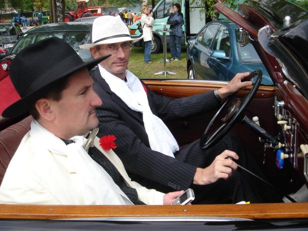 Zwei Männer in Anzügen sitzen in einem Oldtimer-Cabriolet.