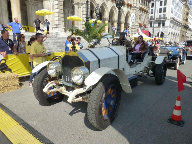 Ein weißes Oldtimer-Auto mit Fahrer und Beifahrerin steht auf einer Straße.