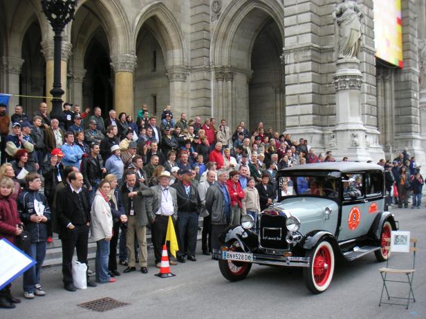 Eine Menschenmenge versammelt sich vor einem Gebäude, während ein Oldtimer im Vordergrund steht.