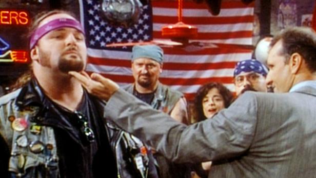 Ein Mann im Anzug berührt den Bart eines Bikers vor einer US-Flagge.