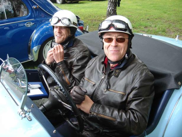 Zwei Männer mit Helmen und Schutzbrillen sitzen in einem Oldtimer.