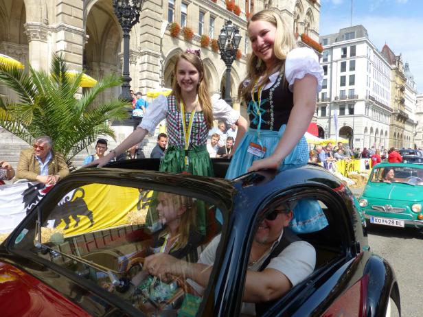 Zwei junge Frauen in Dirndln stehen auf einem Oldtimer in der Stadt.