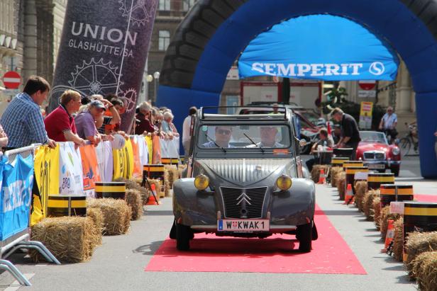 Ein grauer Citroën 2CV fährt auf einem roten Teppich durch eine Stadt.
