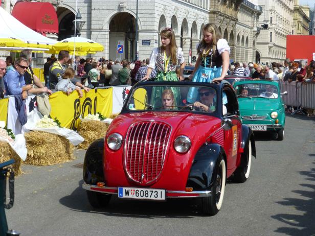 Ein rotes Oldtimer-Cabriolet fährt mit zwei jungen Frauen in Tracht auf einer belebten Straße.