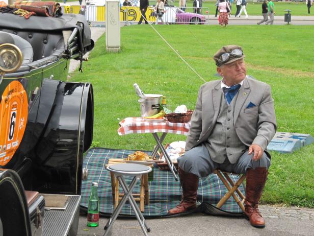 Ein Mann im Tweedanzug macht ein Picknick neben einem Oldtimer.