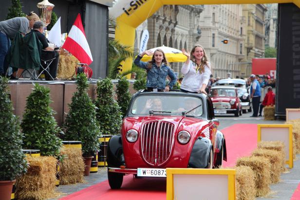 Zwei junge Frauen winken aus einem roten Oldtimer auf einer roten Teppichstraße.