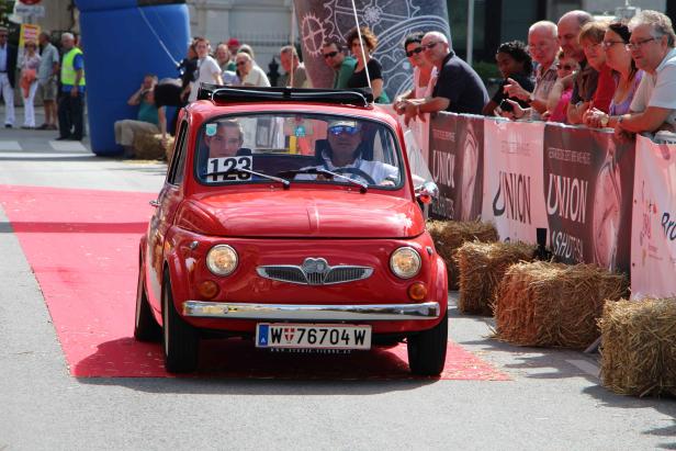 Ein roter Oldtimer fährt auf einem roten Teppich an Zuschauern vorbei.
