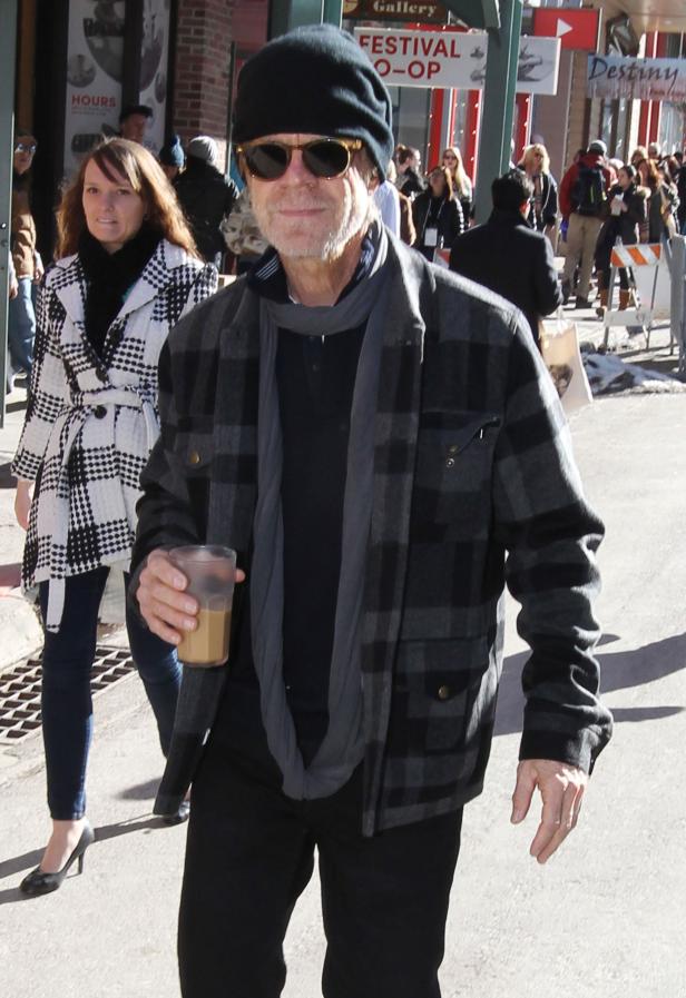 William H. Macy mit Mütze und Schal geht mit einem Getränk in der Hand die Straße entlang.