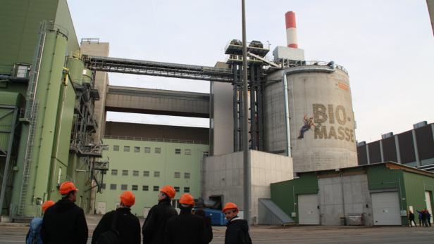 Eine Gruppe von Personen mit Bauhelmen vor einer großen Industrieanlage mit der Aufschrift „Bio-Masse“.