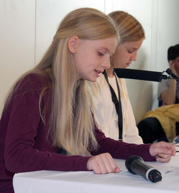 Zwei junge Frauen sitzen an einem Tisch mit Mikrofonen und lesen aus Dokumenten.