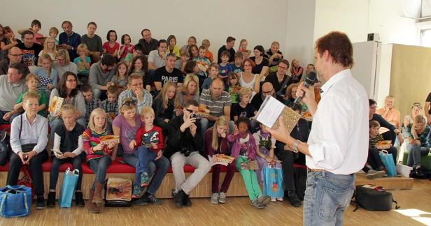 Ein Mann spricht vor einer Gruppe von Kindern und Erwachsenen mit einem Mikrofon in der Hand.