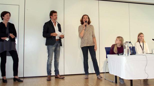 Eine Frau spricht bei einer Podiumsdiskussion in ein Mikrofon, während andere Personen zuhören.