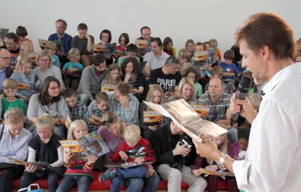 Ein Mann liest vor einer Gruppe von Kindern und Erwachsenen aus einem Buch vor.