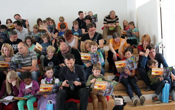 Eine Gruppe von Kindern und Erwachsenen sitzt auf einer Treppe und liest Comics.
