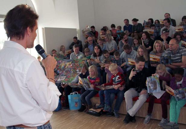 Ein Mann spricht in ein Mikrofon vor einem Publikum, das Bilderbücher betrachtet.