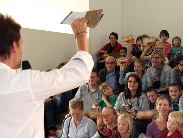 Ein Mann präsentiert ein Blatt Papier vor einem Publikum aus Erwachsenen und Kindern.