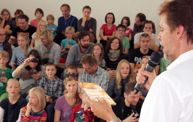 Ein Mann spricht mit einem Mikrofon vor einem Publikum aus Kindern und Erwachsenen.