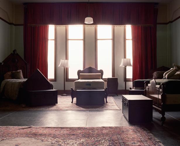 Ein Zimmer mit Betten, Truhen und roten Vorhängen vor hellen Fenstern.